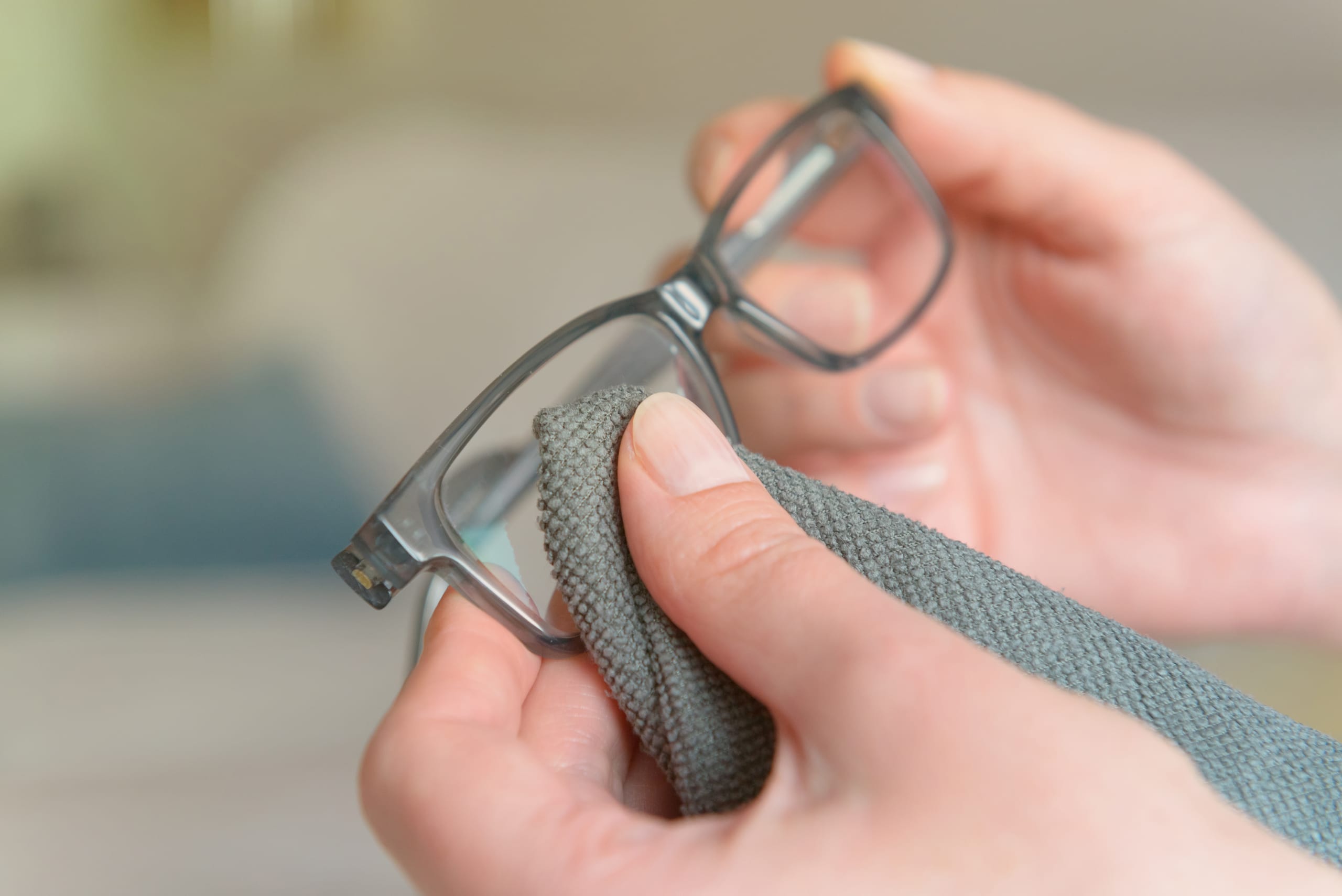 Close-up of hands cleaning a pair of eyeglasses with a gray microfiber cloth. The scene is indoors, with a blurred background.