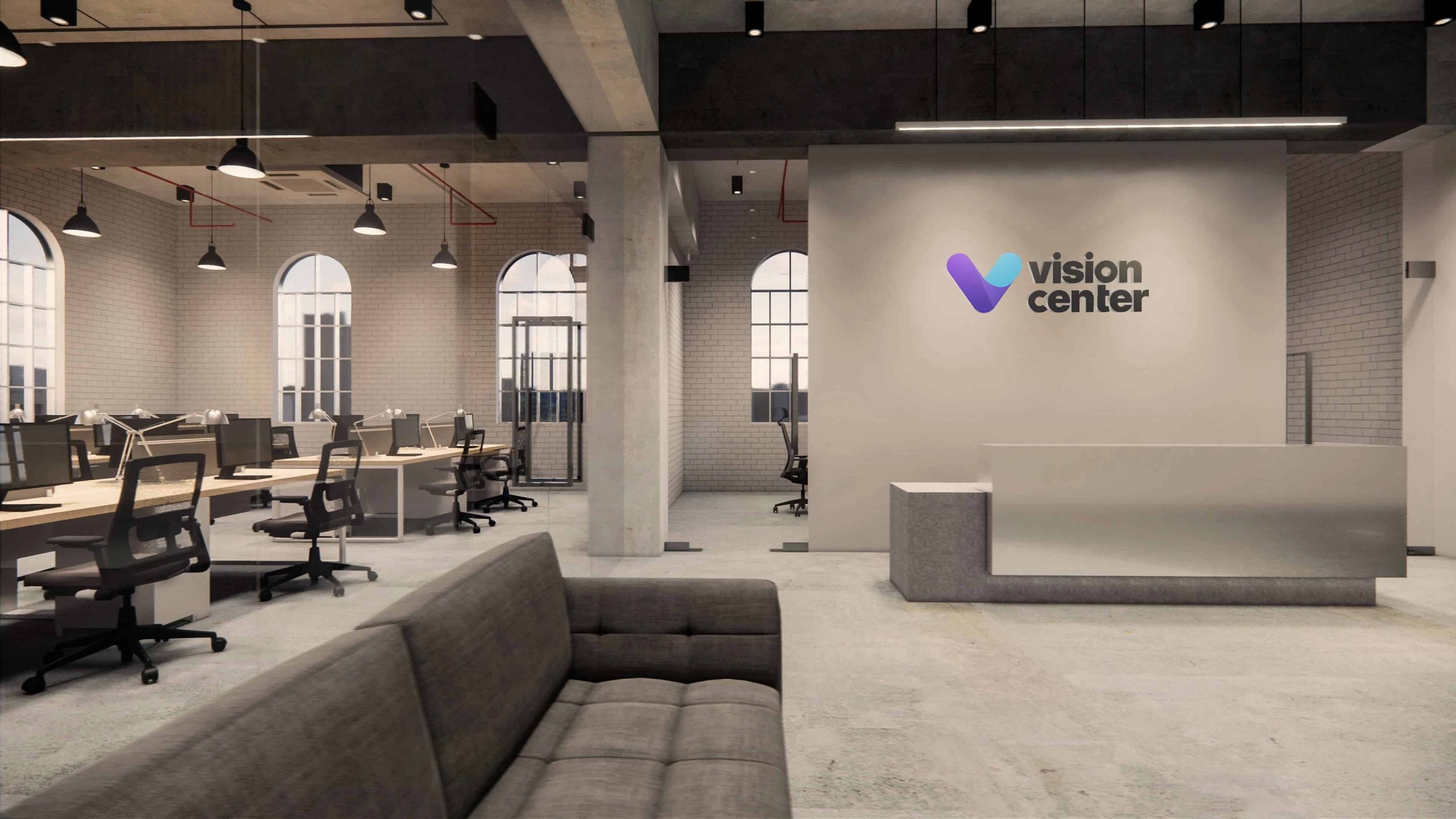 A modern office interior with an open-plan design. There are several desks with chairs, a sofa in the foreground, and a reception area to the right. A sign on the wall reads Vision Center. The space features exposed ceilings and large windows.