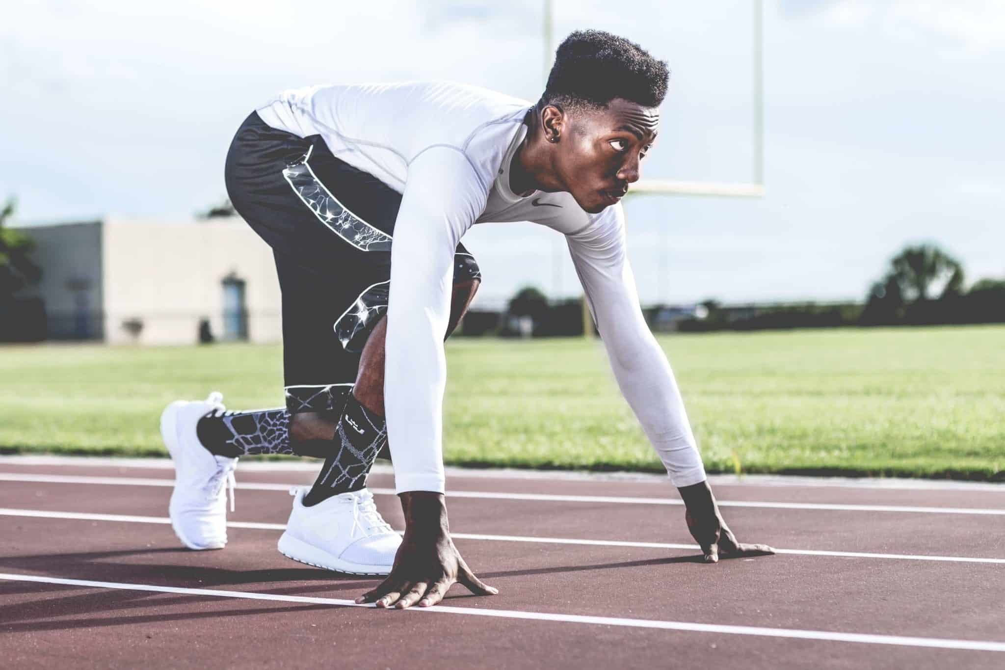 A person in athletic wear is in a starting position on a track, preparing to sprint. The background shows a grassy field and goalposts under a cloudy sky.