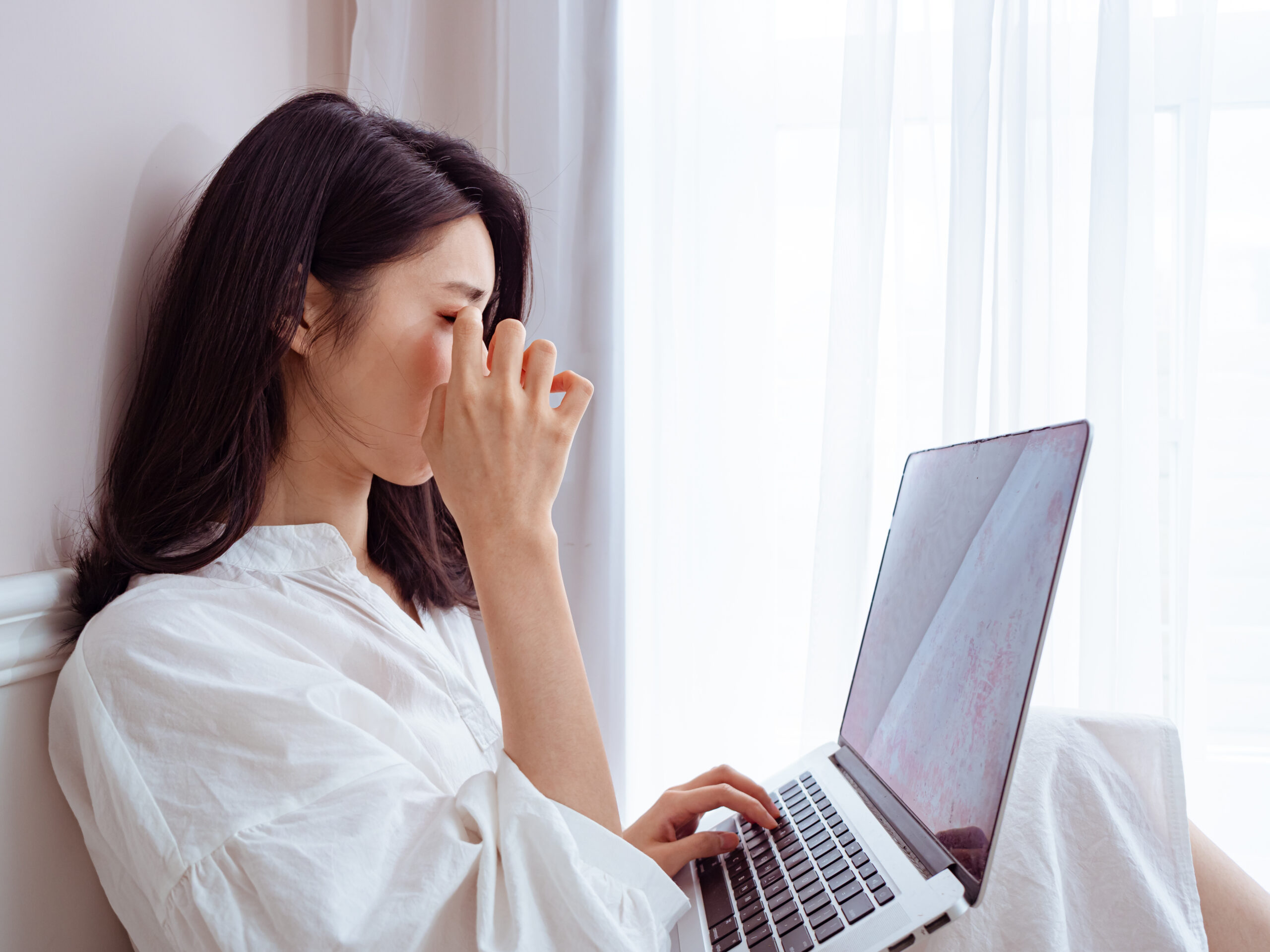 A woman in a white shirt sits by a window with sheer curtains, using a laptop on her lap. She is rubbing her eyes with her right hand, possibly showing signs of fatigue or strain. The lighting is soft and natural.