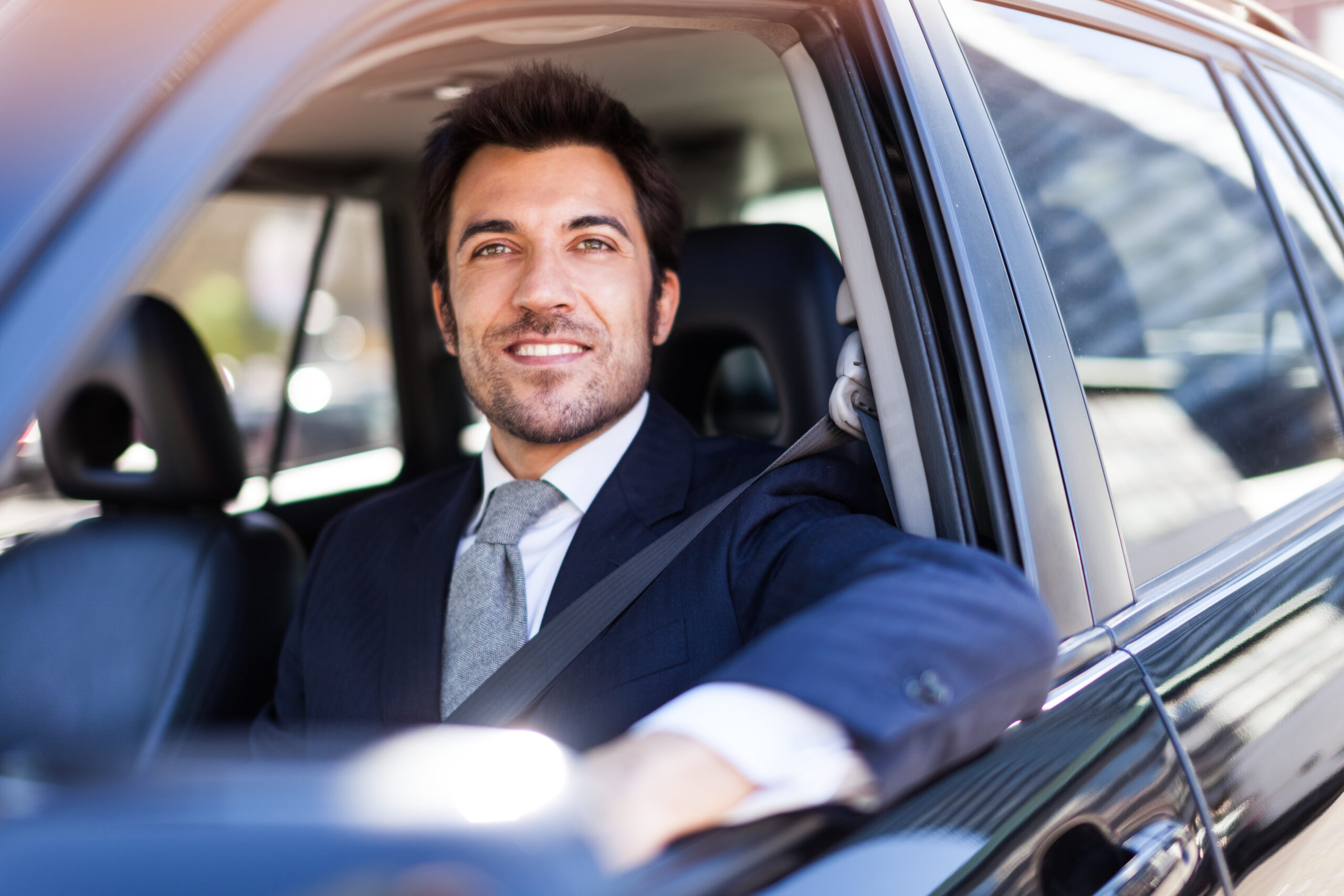 A man in a suit and tie is sitting in the driver's seat of a car with the window down. He is smiling and looking ahead. The interior and part of the exterior of the car are visible.