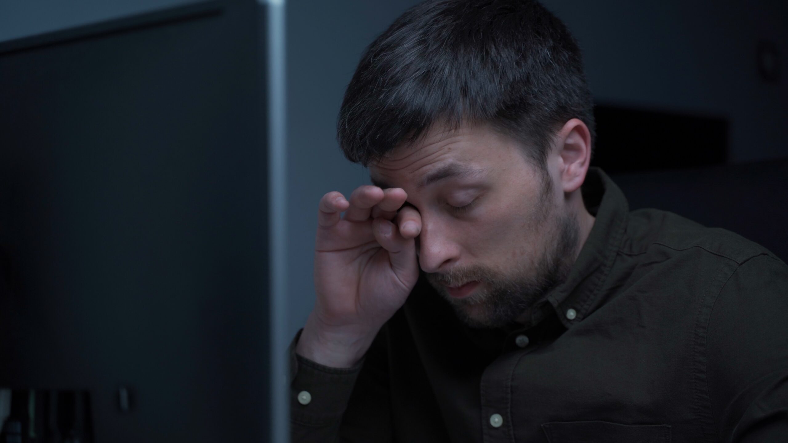 A man with a beard and dark hair wearing a dark shirt sits in front of a computer monitor, rubbing his eye. The lighting is dim, suggesting it might be late or the room is not well-lit.
