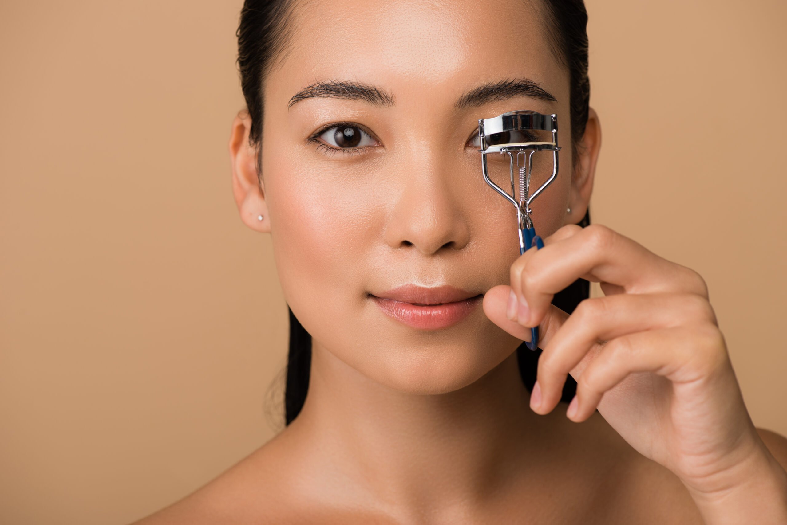 A woman with tied-back hair uses an eyelash curler on her right eye against a neutral background. She appears focused and has a natural makeup look.