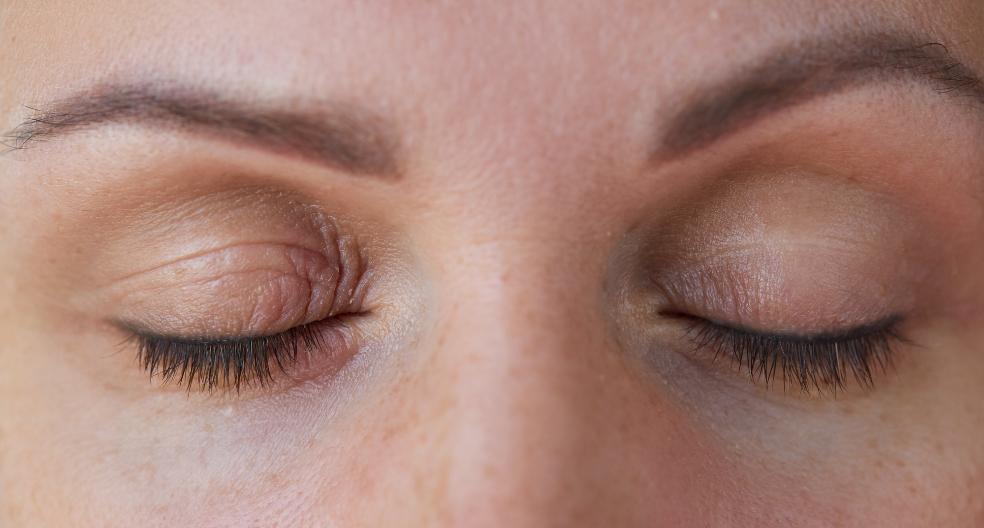 Close-up of a person's face with both eyes closed. The skin around the eyes appears smooth with defined eyebrows. The eyelashes are visible, with light makeup applied to the eyelids. The image is well-lit, showcasing detailed facial features.