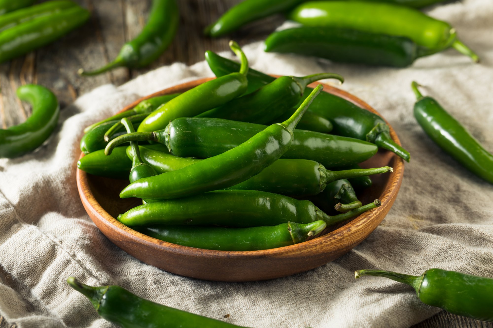 A wooden bowl filled with fresh green chili peppers sits on a textured cloth. Additional chili peppers are scattered around the bowl on a rustic wooden surface.
